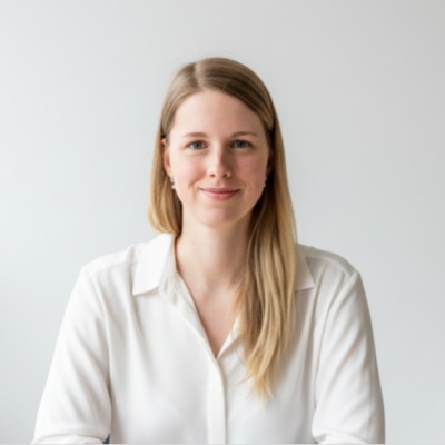 Porträt einer Frau mit langen blonden Haaren und weißer Bluse. Sie blickt mit einem leichten Lächeln direkt in die Kamera vor einem schlichten, hellen Hintergrund.
