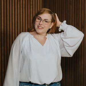 Blonde Frau mit Brille, die lächelt und eine Hand im Haar hat, vor einer Holzpaneelwand.