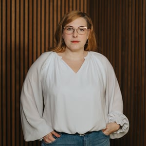 Frau mit blonden Haaren und Brille, in weißem Oberteil, posiert nachdenklich vor Holzpaneelen.