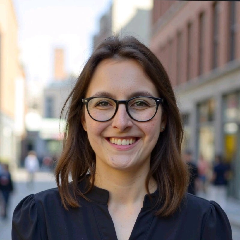 Junge Frau mit Brille, lächelnd, schwarze Bluse aus Blumenstoff, gepflegte Frisur.