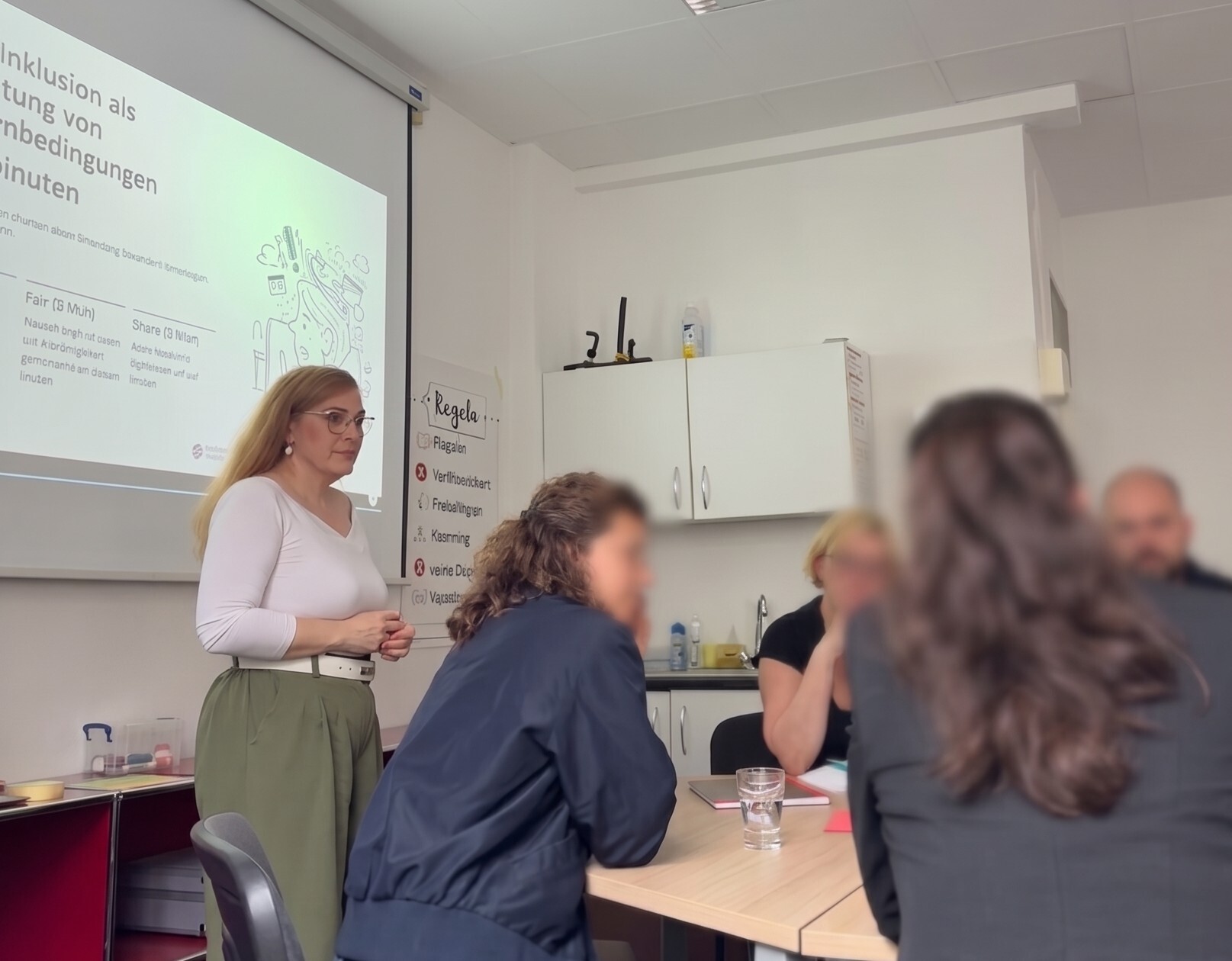 Workshopszene, bei der eine Frau in weißem Obertil und grüner Hose vor einr Leinwand steht und vor ihreine Gruppe von Menschen an einem Tisch sitzt und sich unterhält