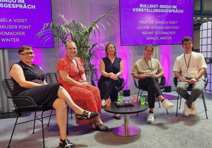 Podiumsdiskussion mit fünf Personen, umgeben von Pflanzen und einem lila Hintergrund.