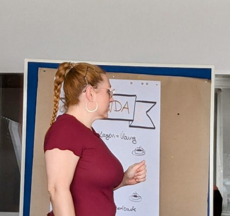 Frau mit langen, lockigen Haaren in rotem Shirt steht vor einer Präsentationstafel.