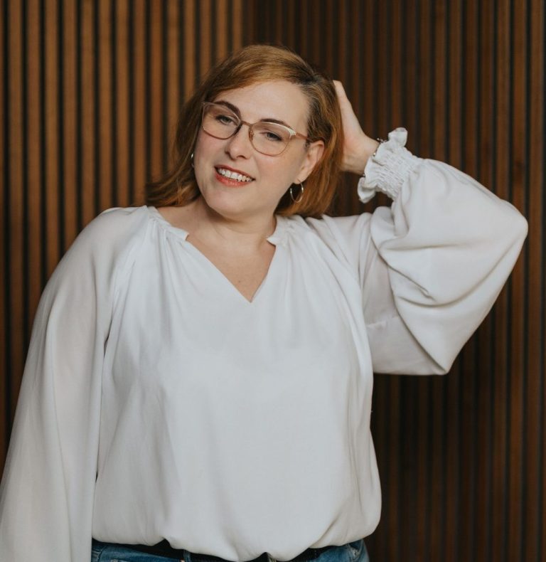 Junge Frau mit Brille, die lächelt und eine Hand im Haar hat, vor einer Holzpaneelwand.