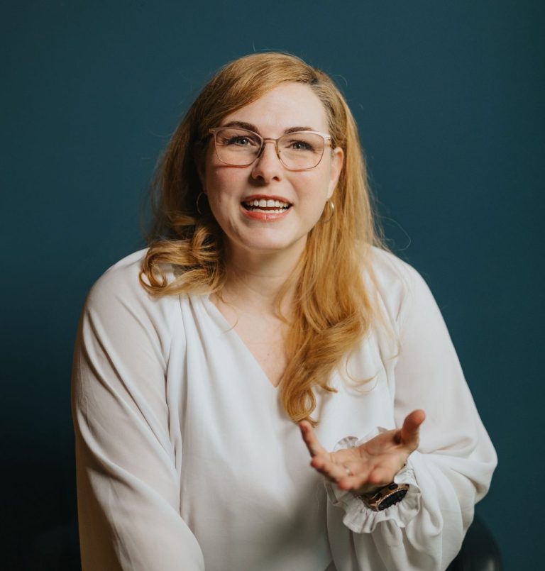 Lächelnde Frau mit blonden Haaren und Brille, die mit einer Hand gestikuliert.